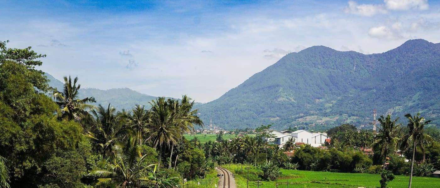 Een spoorlijn loopt rechtdoor tussen groene rijstvelden en palmbomen naar een berg bij Garut
