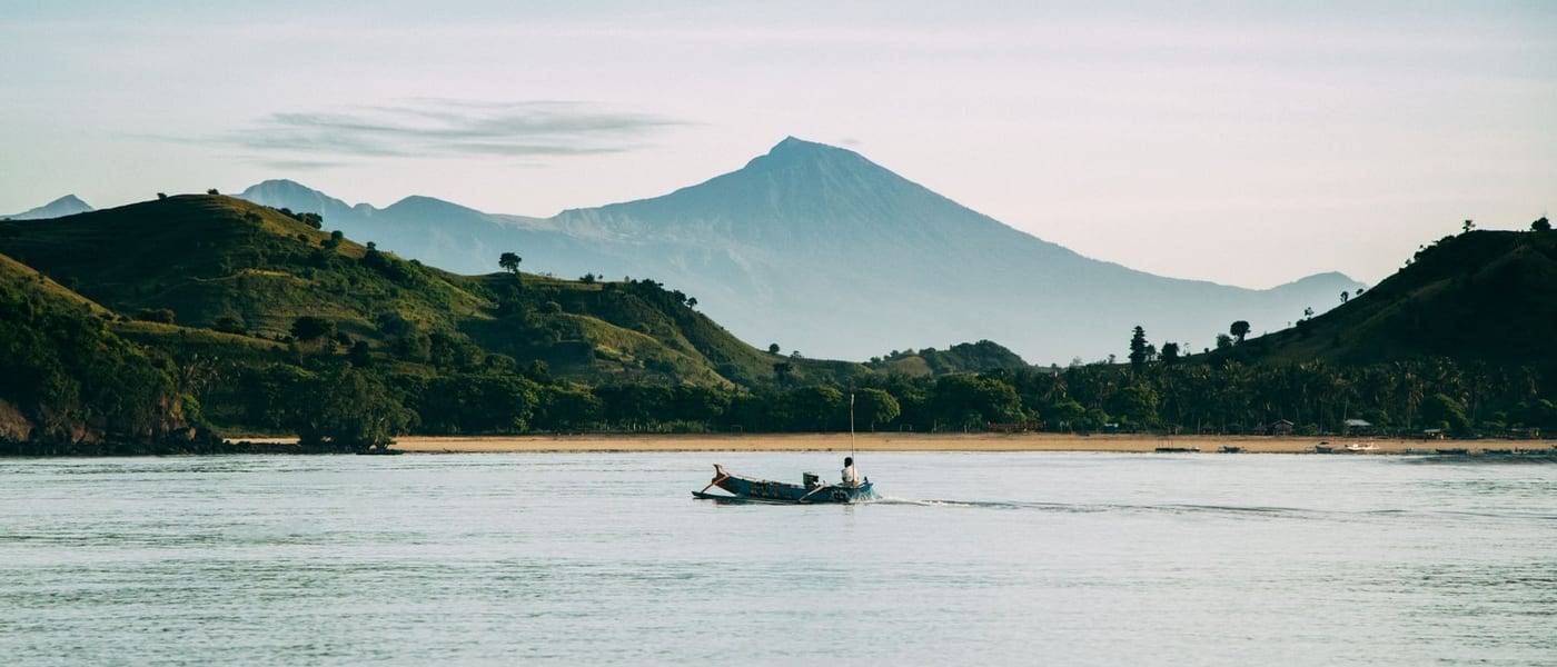 Een vissersboot op kalm water voor groene heuvels, met de Rinjani op de achtergrond